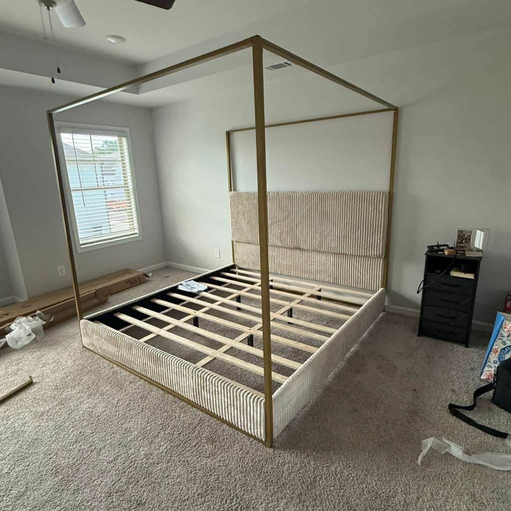 Upholstered canopy bed frame with gold-tone posts and exposed support slats during assembly.